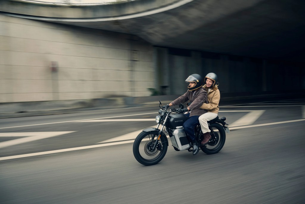 A couple riding on a Maeving RM2 on a road with a curved overpass above.