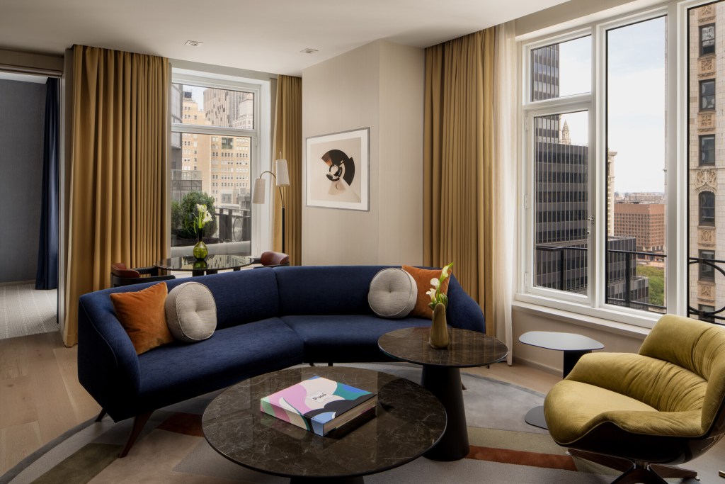 A guest room at Four Seasons New York Downtown with a blue curved sofa, neutral three round coffee tables, a mustard chair and views of Tribeca outside the windows.