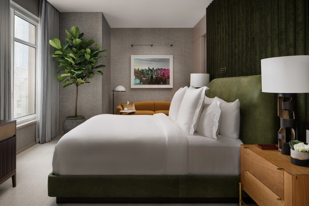 A guest room at Four Seasons New York Downtown with a white bed facing a window, potted tree in the corner and a cityscape photograph on the wall.