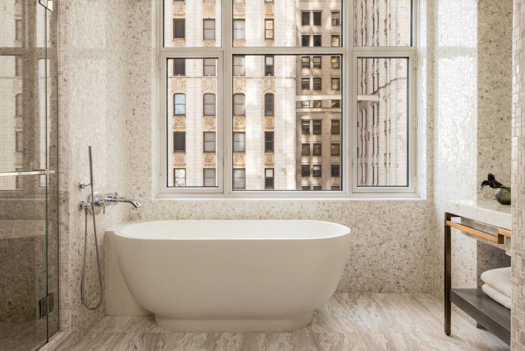 A white soaking tub in the bathroom at Four Seasons New York Downtown with small neutral-colored tiles on the floor and wall and a large window with views of Tribeca buildings outside.