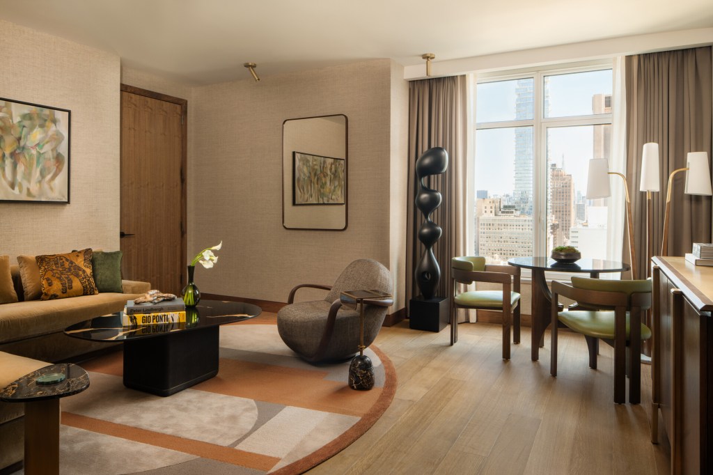 A guest suite at the Four Seasons New York Downtown with a large window on the right, a sofa and chair with a round rug by the door and a small table with chairs and a tall sculpture in front of the window.