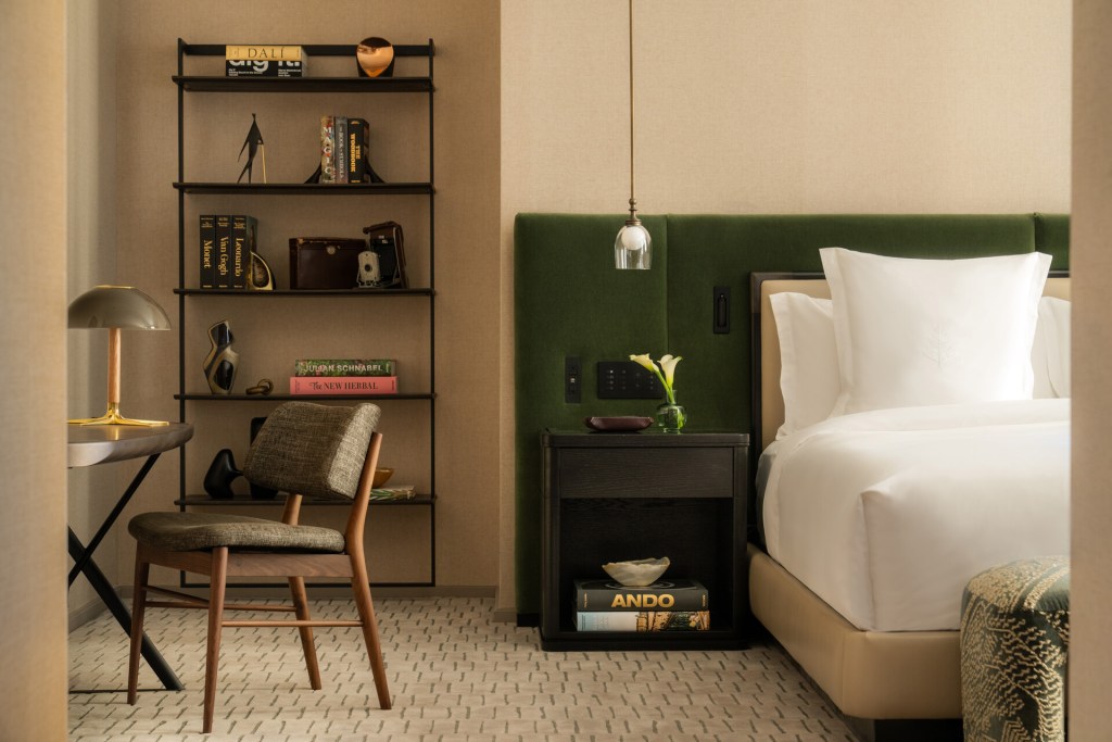 A guest room at Four Seasons New York Downtown with a bed with a dark green headboard, a shelf to the left with art objects and books, and a small table with a wooden chair.