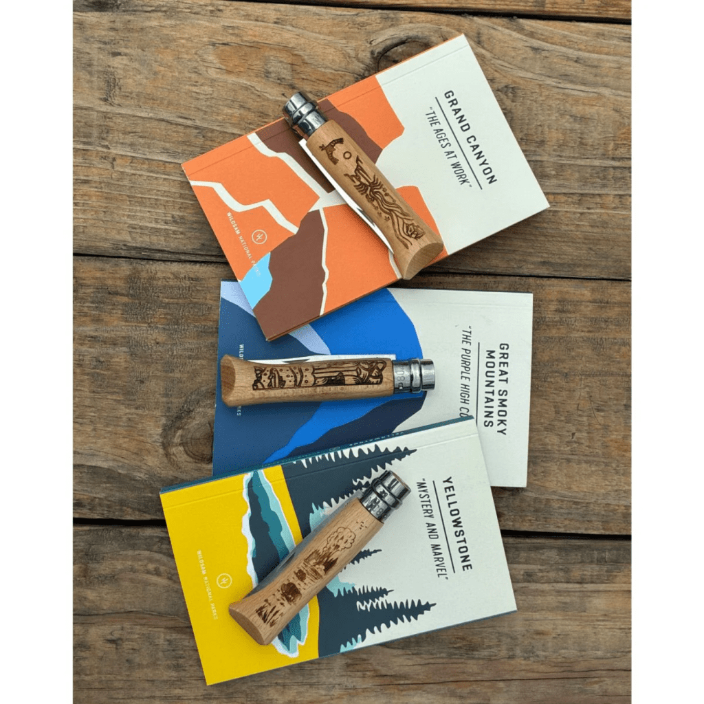 Three national park Opinel knives laying on top of their corresponding Wildsam National Park guide on a wood plank surface.