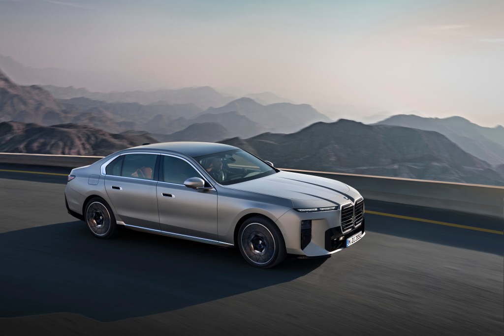 A silver BMW 7 Series driving on a windy road with mountains and clouds in the background.