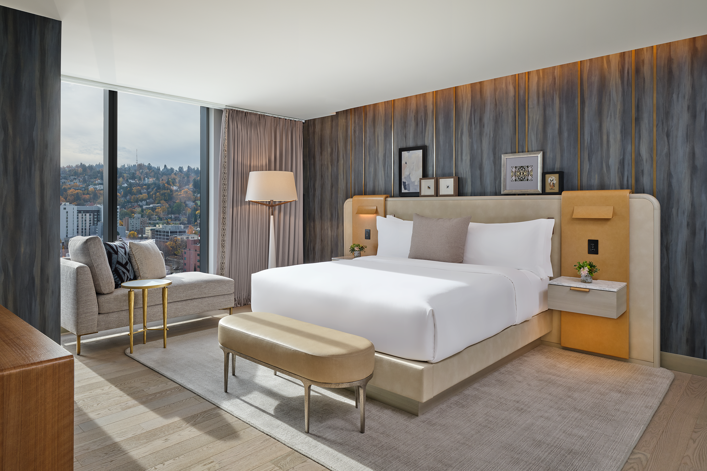 A guest room at The Ritz-Carlton, Portland with white bedding, a tan bench and nature-inspired wallpaper, looking out onto a large window with a view of the hills.