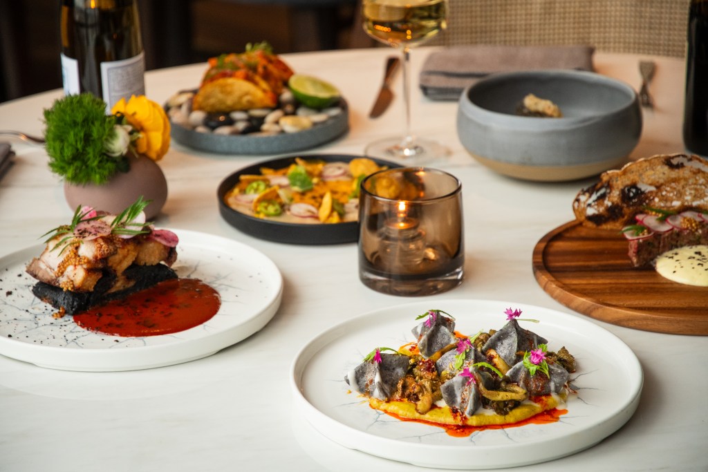 A collection of several plated dishes, a glass of wine, a bowl of apps and a candle in a gray glass holder at Bellpine.