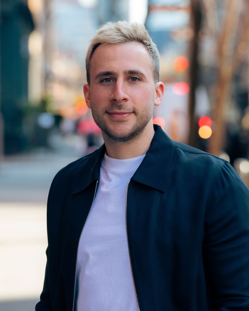 A portrait of Rumor founder Josh Zipkowitz, blonde haired man wearing a navy zip-up jacket and white tee shirt, with a soft focus street behind him.