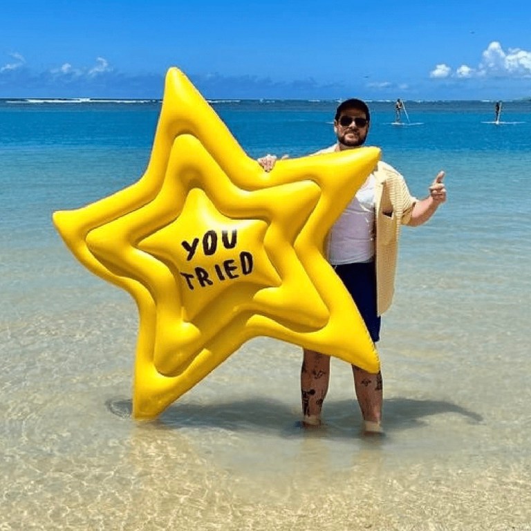 A man standing in the ocean holds the "You Tried" inflatable pool float by Adam JK