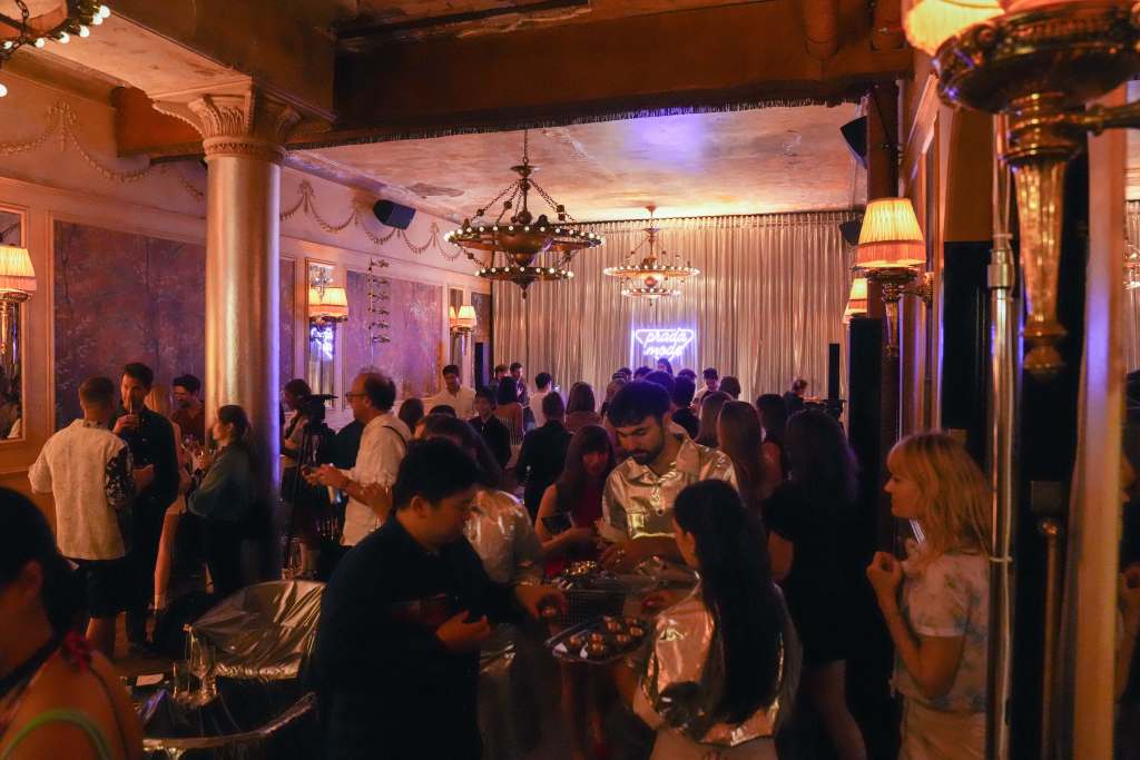 A crowd of people gathering in a dimly lit room, in front of a small stage with a silver curtain and a purple neon sign that reads "Prada Mode."