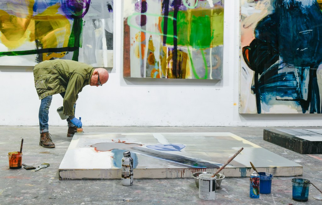 Aurel K. Basedow leaning over a large canvas on the floor and pouring epoxy on it.