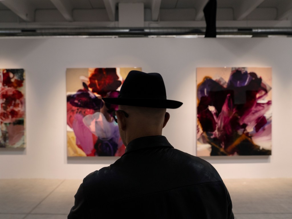 Aurel K. Basedow, shot from the back, wearing a brimmed hat and looking at colorful abstract artworks hanging on the wall in front of him.
