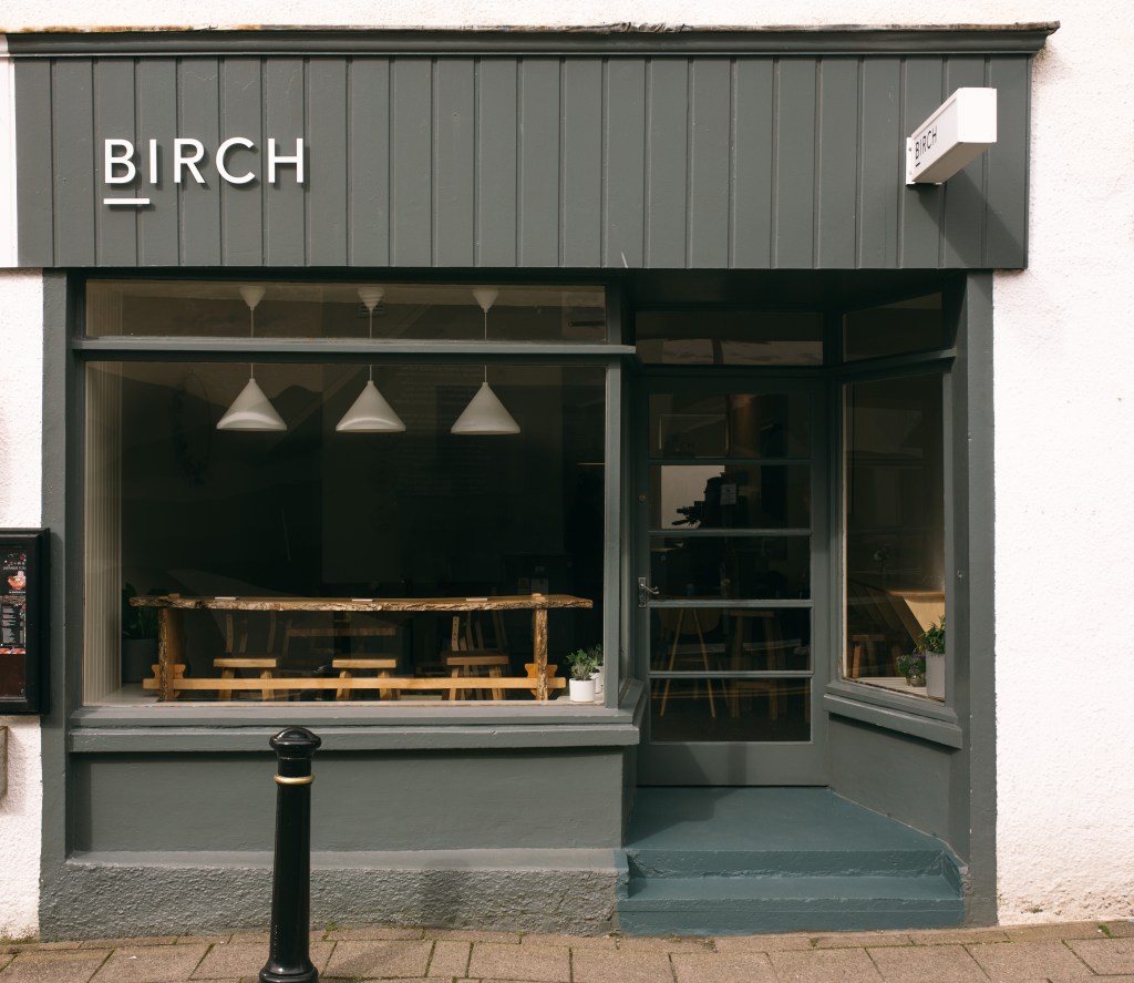 An exterior shot of a gray coffee shop that a minimal sign that syas "Birch" at the top left of the facade.