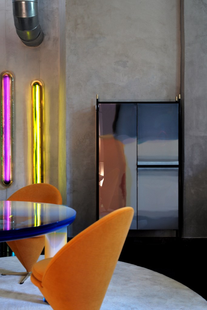 Two orange chairs sitting at a blue translucent table with a colorful light installation and a mirrored cabinet behind it.