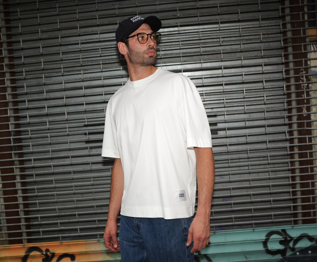 A model wearing a white RANDOM/RANDOM t-shirt, standing in front of a silver store gate and looking away from camera.