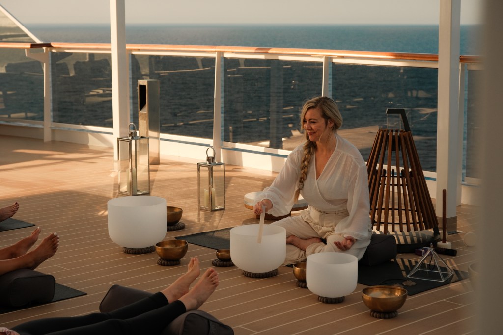 A woman with long blonde hair sits on the deck of a ship with the ocean in the background and sound bowls in front of her.