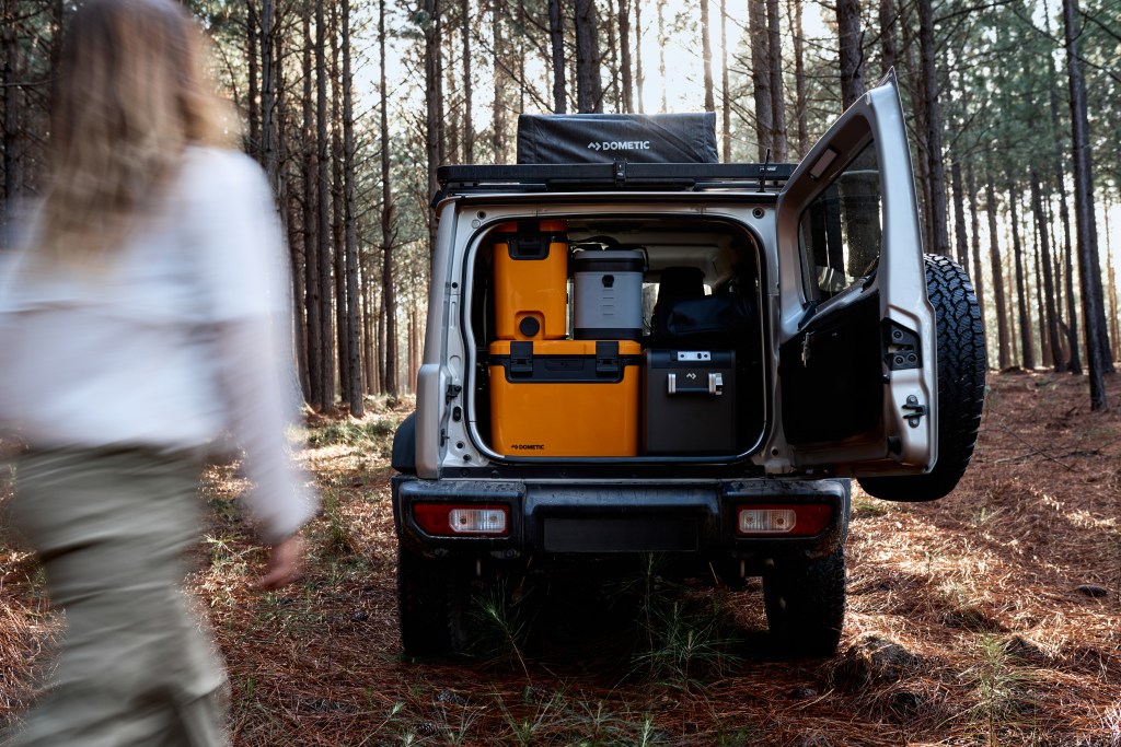 A shot of the back of an SUV with the hatch open, revealing a collection of gray and orange Dometic coolers, stacked in the back with a person walking towards it.
