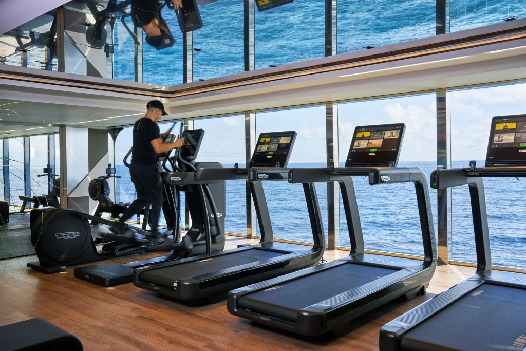 A man running on a Technogym elliptical in a gym room with floor-to-ceiling windows overlooking the ocean.