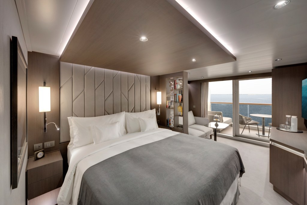 An interior view of the Ocean Terrace Suite showing a large bed with gray linens and a sliding glass door that opens up to a private terrace overlooking the ocean.
