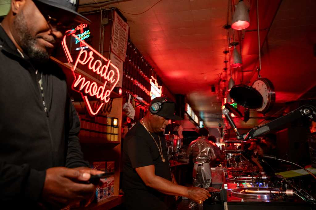 Grandmaster Flash and another man in the foregound playing records at a crowded, red-lit room with a red neon sign behind them that reads "Prada Mode."