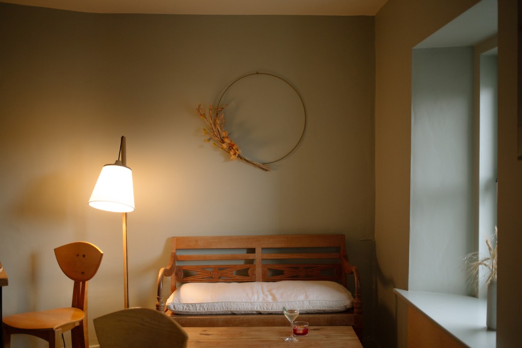 A small room with a light wood bench, a glass of wine and a cocktail on a table and a window on the right side of frame.