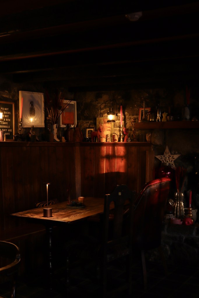 A bar corner with a wood table, brick walls and a variety of framed art with warm, moody light. 