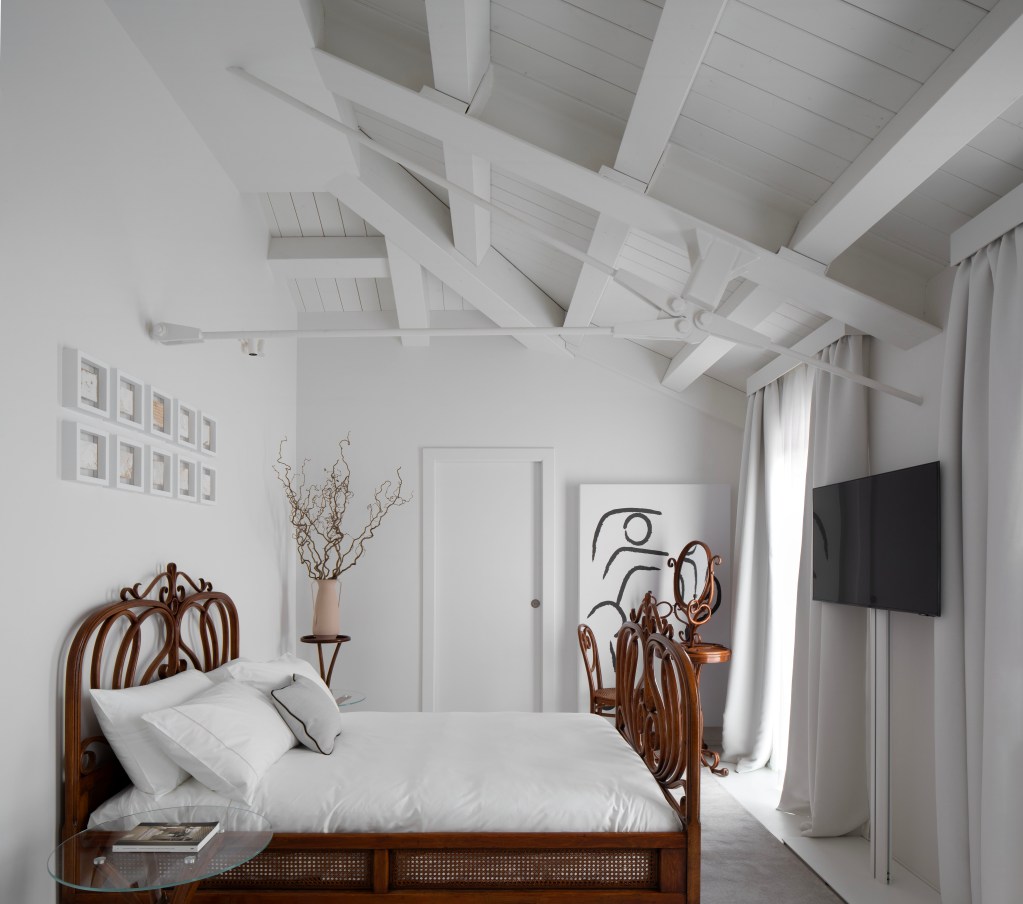 The all-white Thonet room at Interno Marche with a tall ceiling, white walls, long curtains, minimal art and a brown wood and cane bed.