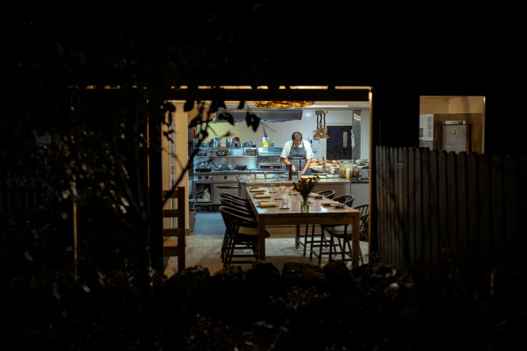 A view through a window of a chef prepping food in a restaurant kitchen with a long dining table in front. 