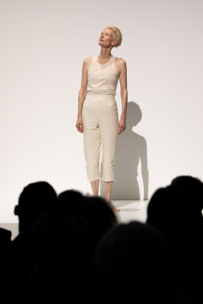 Tilda Swinton wearing a cream tank top and pants, standing and looking up to the left with a seated audience in the foreground.