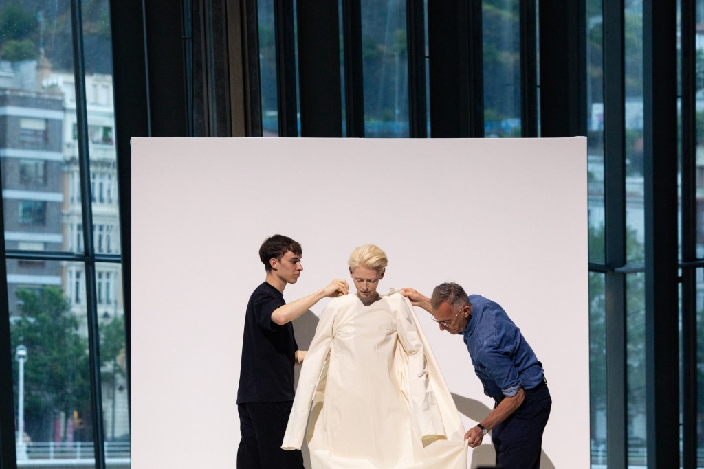Tilda Swinton standing in between and two actors who are changing her all-cream outfit as part of her performance art piece.