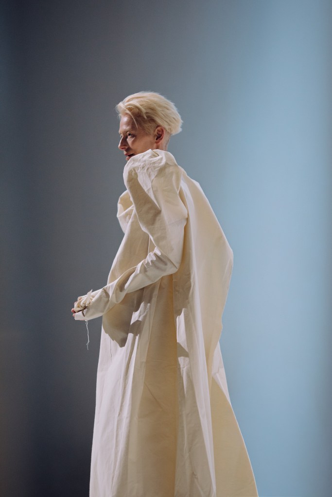 Tilda Swinton wearing a long cream dress with a blue-ish gray background behind her, looking off camera to the left of frame.