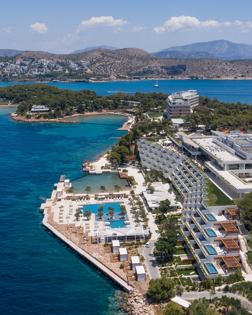 An aerial shot of the Four Seasons Astir Palace Athens property on a peninsula surrounded by turquoise blue water, green trees and mountains in the distance.