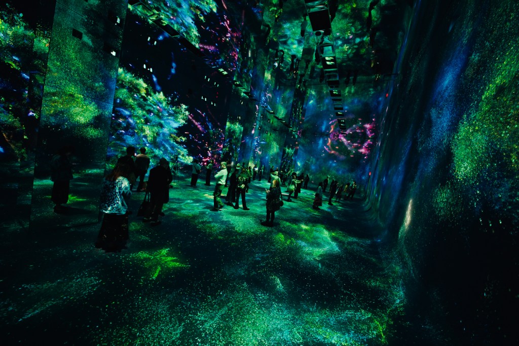 A group of people in a large gallery room with blue, green and black digital art projected on the ceiling, walls and floor.