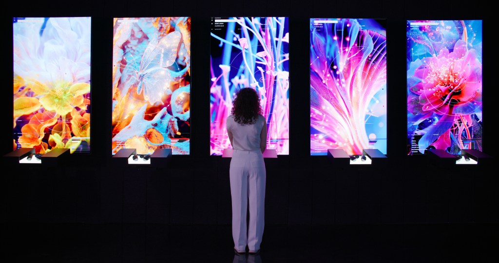 A woman wearing all white stands in front of five vertical AI art screens at Dataland.
