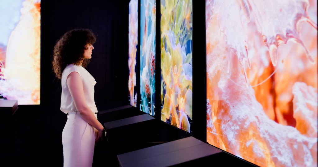 A woman with dark curly hair wearing all white looks at large panels of AI art.