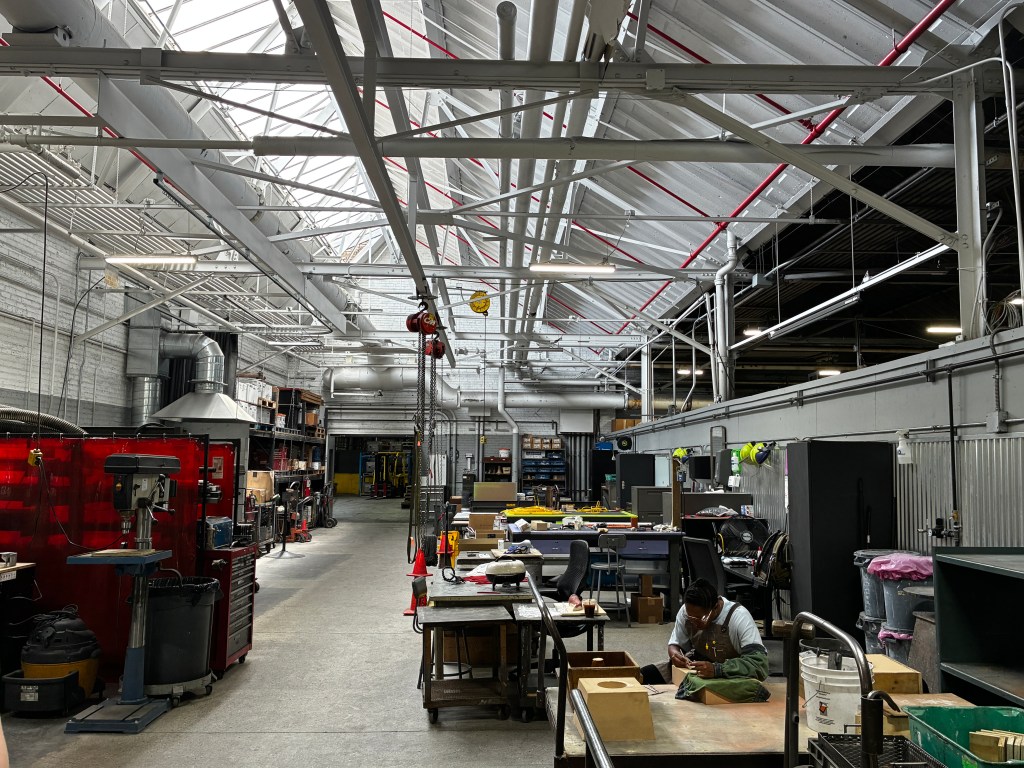 A long industrial room with tall ceilings flanked by studio spaces and foundry supplies with an artist working on the right side of the space.