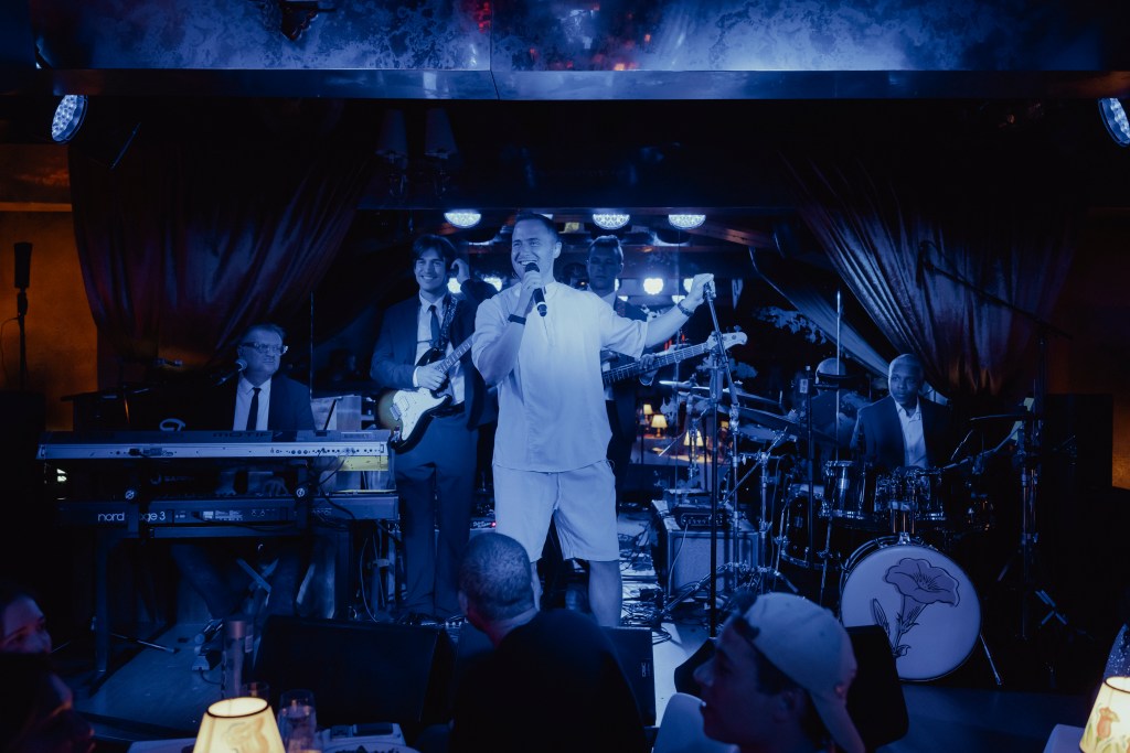 Mike Posner on a stage lit up in blue lights, standing in front of a crowd with a band behind him.