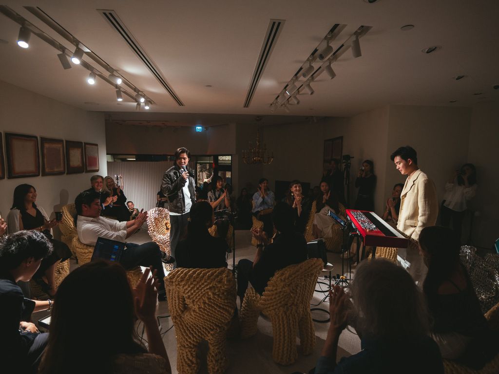 A group of people sitting in a dark room with a person standing at the front of the group about to play on a red keyboard.
