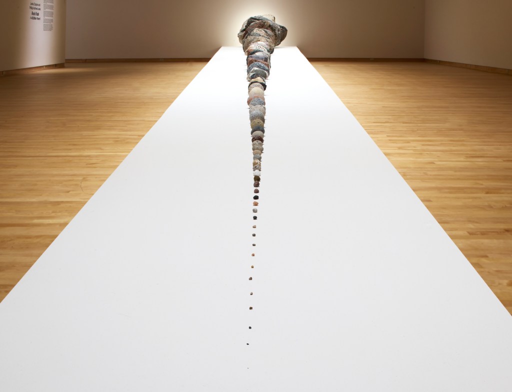 Lenka Clayton and Phillip Andrew Lewis's Rock Fade (≈ 45 Billion Years), shot from the smallest rock looking towards the largest rock of the installation, sitting on a long white platform with a light wood floor background.