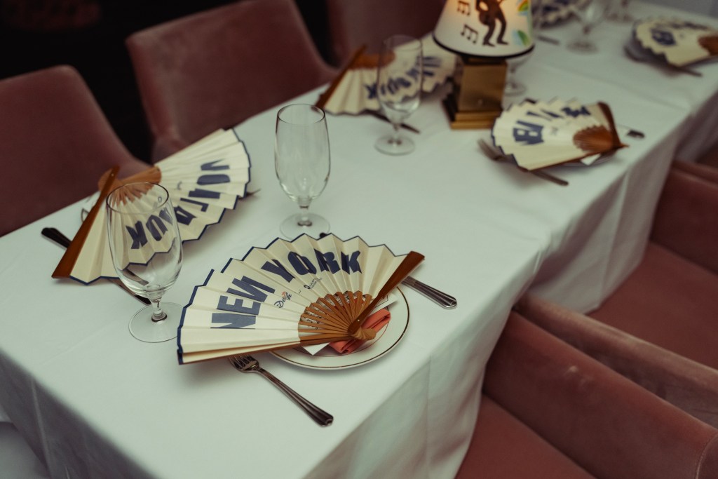 A table with place settings, plush pink chairs and paper fans with "NEW YORK" printed on them.