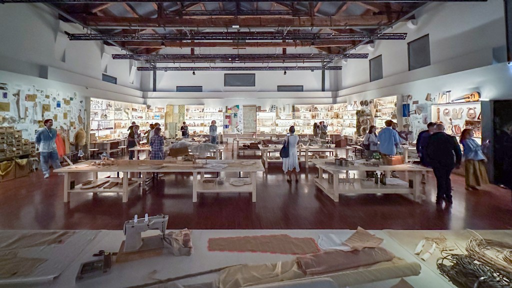 A large room made to look like an atelier, with illuminated shelving and large wooden tables of artisan supplies and artworks.