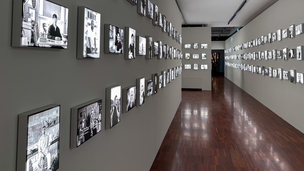 A hallway full of framed black and white photographs of artisans hanging on both walls with a dark floor leading to a doorway.