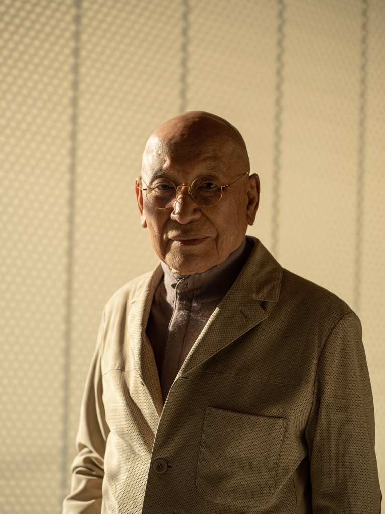 A portrait of Park Seo-Bo in warm light, wearing a tan patterned jacket with a high-collared sweater underneath.