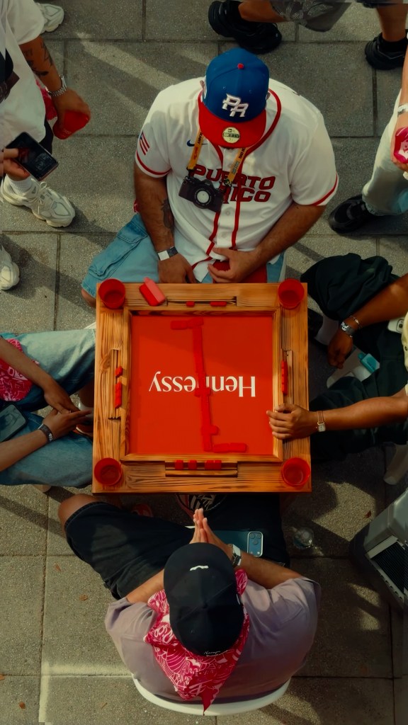 An overhead shot of four men playing dominoes on a red table that says "Hennessy" on it.