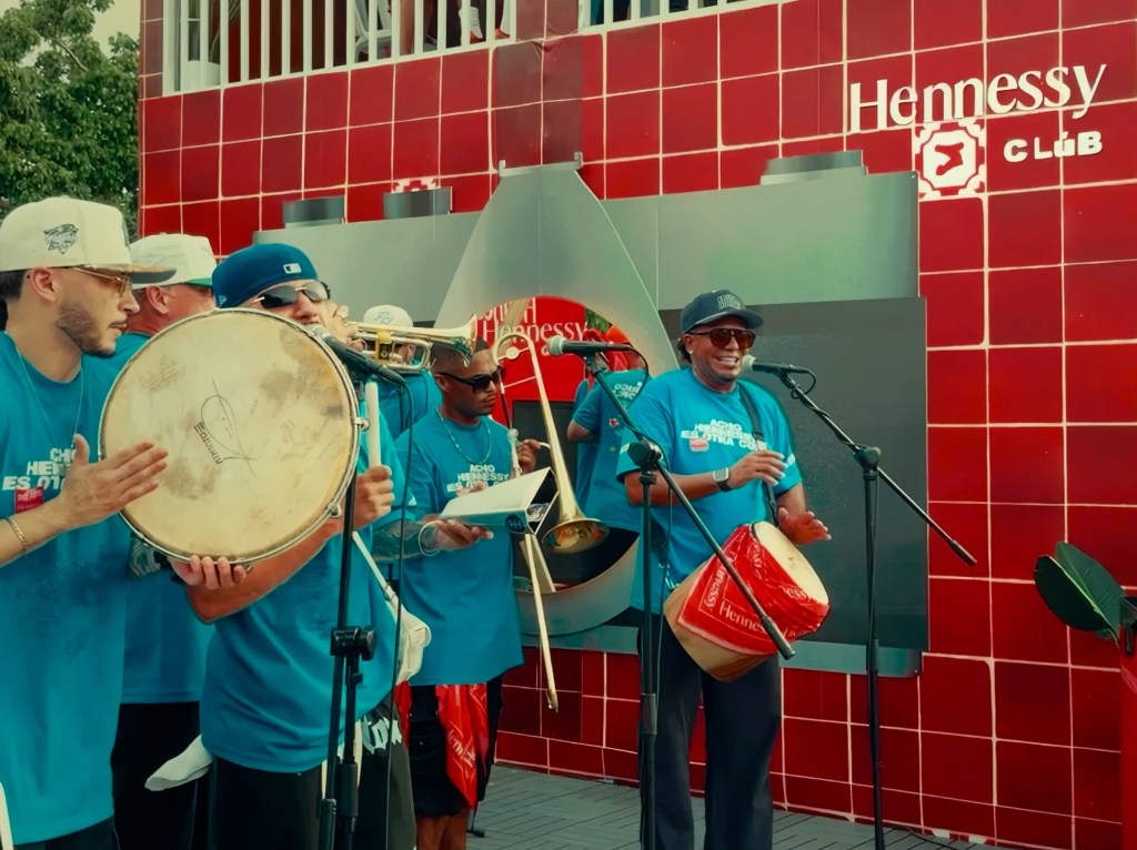 A band of musicians wearing teal shirts play instruments in front of a red tiled wall that says "Hennessy Clúb."