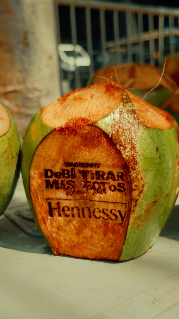 A coconut with a cut side and the words "Bad Bunny DeBÍ TiRAR MáS FOToS World Tour Hennessy" burnished into its flesh.