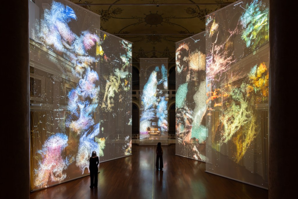 Several large vertical screens with images projected on them as people stand below looking up at them.