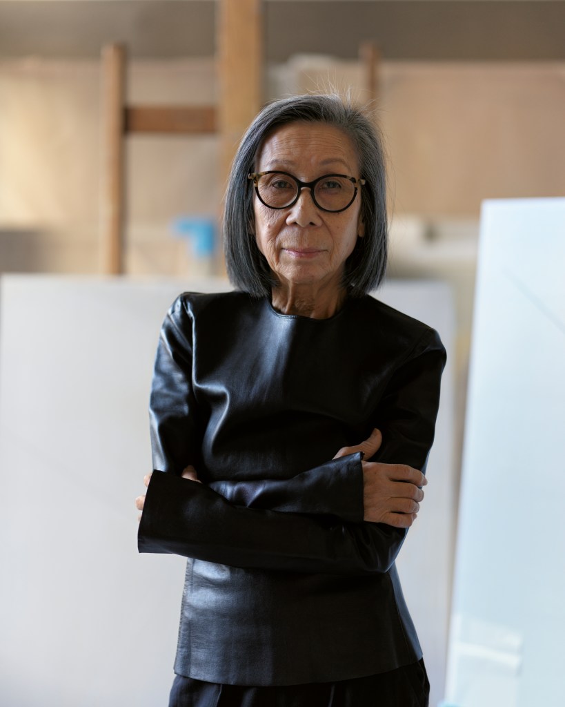 A portrait of Huong Dodinh, standing with her arms crossed in a large studio space with artworks behind her.