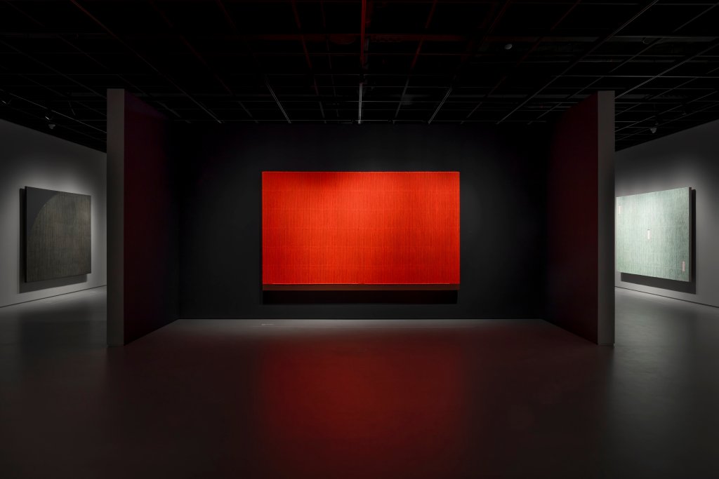 A horizontal, bright red-orange work by Park Seo-Bo, displayed on a black wall with other works on nearby walls.
