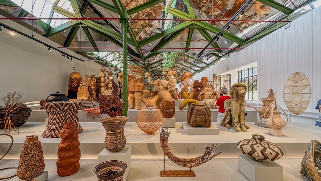 A room with light wood platforms and a large green latticed ceiling with a series of woven objects and baskets displayed in rows.