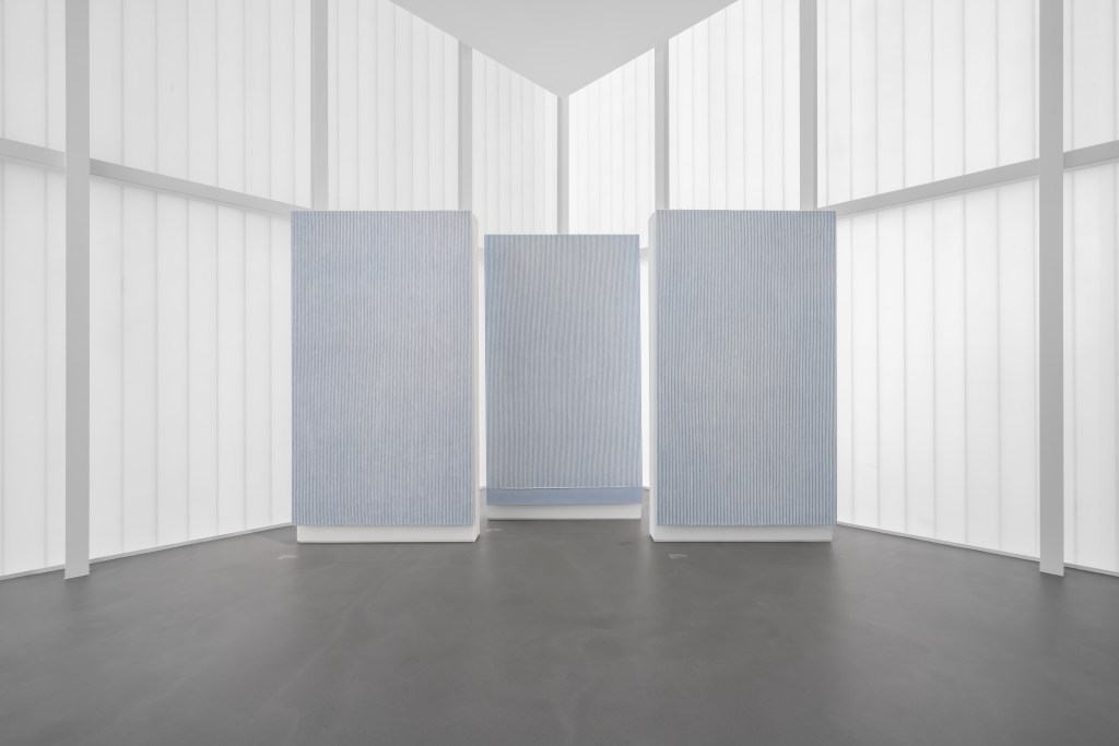 An installation view of three vertical artworks displayed as a trio in a white, gridded room and dark gray concrete floor.